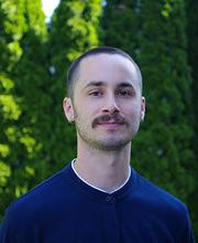 Headshot photo of Professor Eric Aldieri with very short brown hair and mustache wearing navy blue jersey with white trim collar in front of tall green bushes