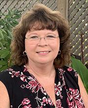 headshot photo of Dr. Andrea Cayson with medium length brown wavy hair wearing clear rimmed glasses and a sleeveless v-neck black blouse with pink floral print