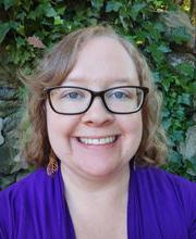 Dr. Ashey Hansen-Brown smiling with medium length wavy light brown hair and wearing brown rim glasses and a royal blue v-neck top