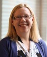 Dr. Heather Marella smiling with medium length blonde hair wearing brown rim glasses and a vneck 黑色的 white and purple print dress with a purple jacket