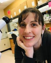 Dr. 乔斯林米尔斯 next to microscope in lab smiling with long dark brown hair pulled back in a pony tail with bangs and wearing earphones and a 黑色的 long sleeve top