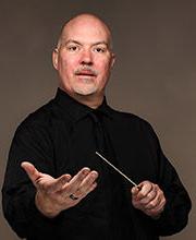 Dr. Donald Running wearing a black button down shirt with black tie, holding a conductor's wand in one hand while his other hand is held out towards the camera