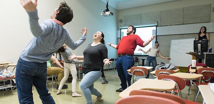 an acting class with the students walking in a circle with their arms out wide, leaning back and yelling while the teacher directs from the front corner of the room