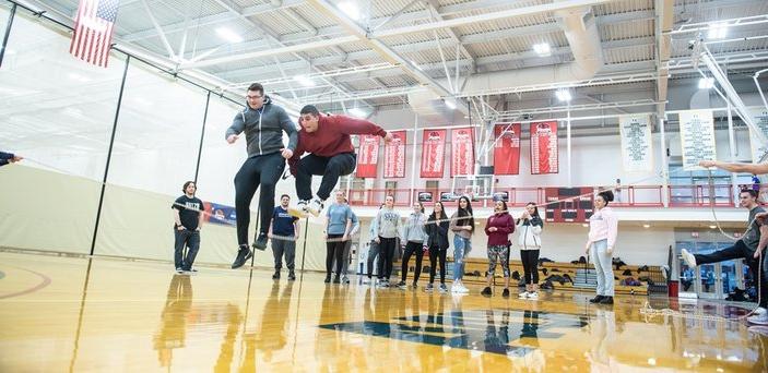 2 male students jumping over a rope in Physical Education class in a gymnasium as other students cheer them on